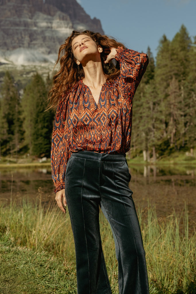 Woman in a red and blue print blouse standing in a natural setting with trees and mountains.