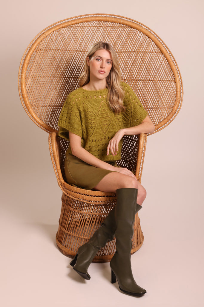 Sitting shot of model wearing a green knitted short sleeve relaxed fit jumper in a brown wicker chair.