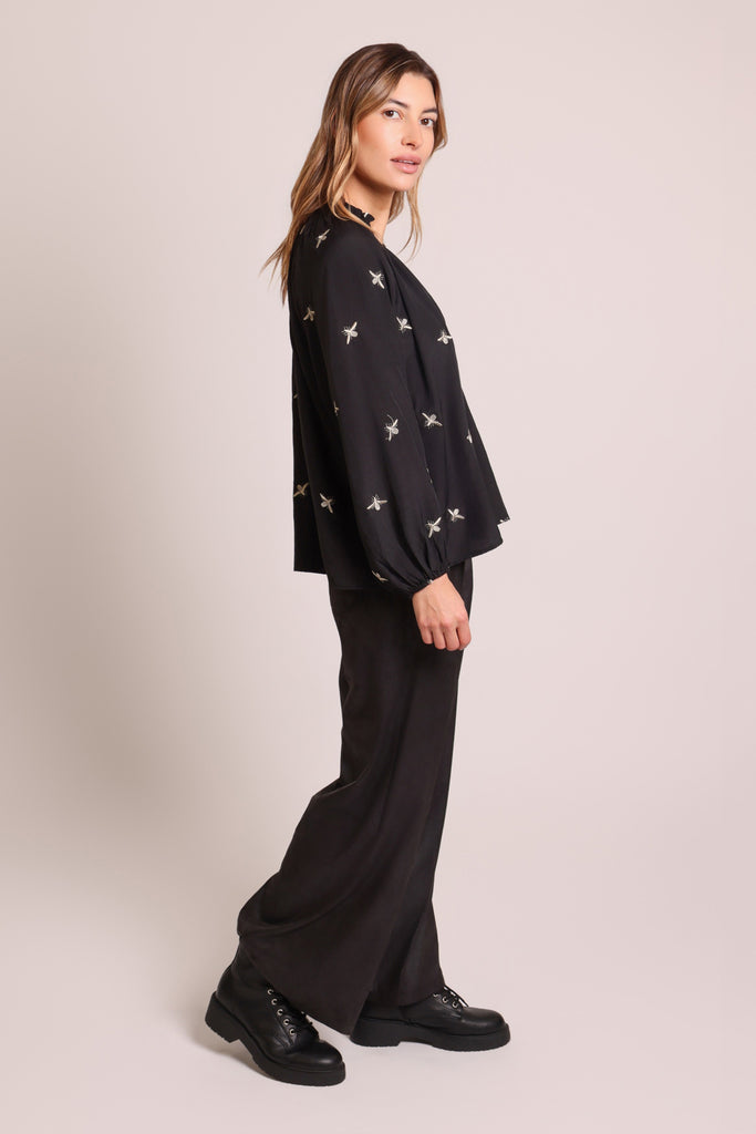 Side shot of model wearing a black long sleeve V-neck relaxed fit blouse with metallic bee embroideries.