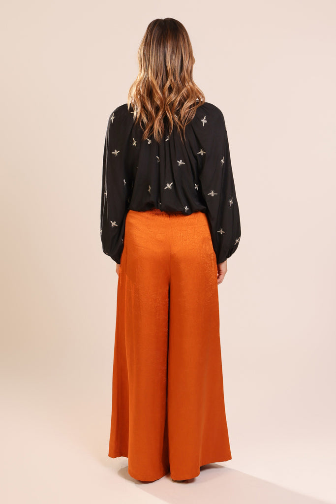 Back shot of model wearing a black long sleeve V-neck relaxed fit blouse with metallic bee embroideries.
